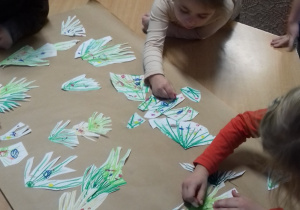 grupa dzieci przy stole przykleja wycięte elementy choinek na szarym papierze